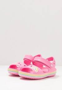 Vaaleanpunaiset sandaalit, joissa on avoimet varpaat ja jotka on valmistettu kumista. Niissä on monivärinen sateenkaari-yksityiskohta ja raidallinen pohja pasteliväreissä. Tarrasuljenta.