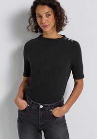 Kvinde iført ribbet sort t-shirt med tre sølvknapper på den ene skulder, stoppet ned i højtaljede sorte jeans, hænder i lommerne.