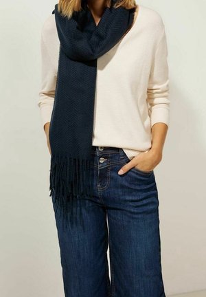 Femme portant un pull crème, une écharpe tricotée bleu foncé avec des franges, et un jean bleu, une main dans la poche.