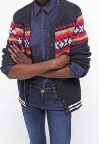 Cárdigan de punto grueso en gris oscuro con franjas horizontales multicolores. Presenta un cierre de botones y acentos a rayas blancas en el dobladillo.