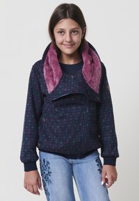 Ragazza che indossa una giacca navy a fantasia con un grande colletto rosa morbido e jeans azzurri chiari con ricami floreali, in piedi contro uno sfondo semplice.