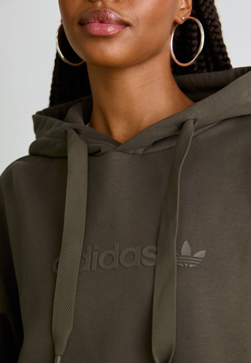 Olivgrön hoodie med dragsko, som har en framträdande präglad logotyp. Tillverkad av ett texturerat tyg med en mjuk, avslappnad design.