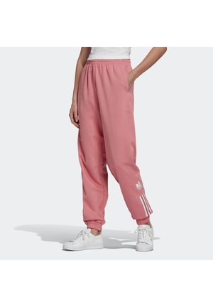 Persoon die roze Adidas trainingsbroek draagt met witte strepen en een logo bij de enkel, gecombineerd met witte sneakers en een wit shirt.