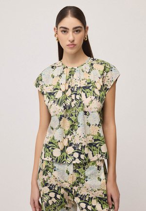 Donna che indossa top e pantaloni floreali abbinati con fiori verdi, beige e blu, in piedi davanti a uno sfondo chiaro uniforme.