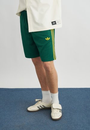 Persona che indossa pantaloncini verdi Adidas con strisce gialle e logo, calzini bianchi, scarpe da ginnastica bianche Adidas, in piedi su tappeto blu.
