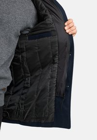 Marinblå jacka med dragkedja fram och kviltad svart foder. Har en innerv pocket och mjuk texturerad utsida. Öppen och delvis på.