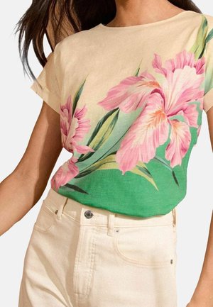 Frau trägt ein beigefarbenes Kurzarmoberteil mit großem pinkfarbenem Blumenmuster und hochtaillierte, cremeweiße Hose.