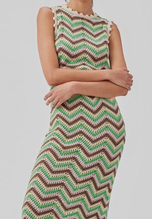 Femme portant une robe en crochet multicolore sans manches avec des motifs en zigzag verts, marron, roses et crème, les bras croisés sur le torse.