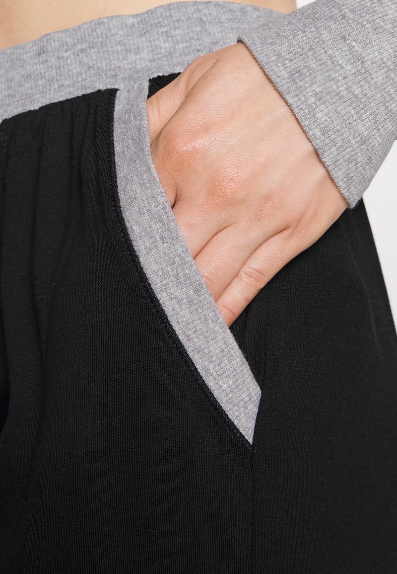 Pantalones negros con una cintura y acentos de bolsillo en gris, que presentan una tela suave y elástica con un ajuste relajado. Mano colocada en un bolsillo.