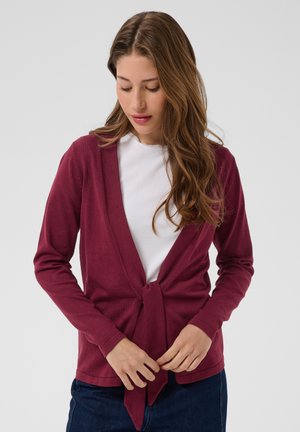 Jeune femme faisant un nœud à l'avant d'un cardigan bordeaux par-dessus un top blanc, portant un jean bleu foncé, sur un fond clair uni.
