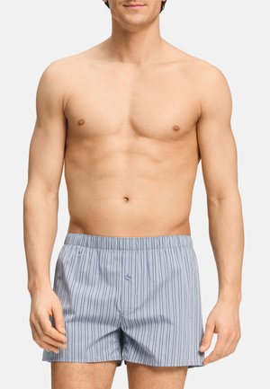 Hombre sin camiseta que lleva puestos unos calzoncillos tipo bóxer de color gris claro con finas rayas negras verticales y un botón delantero, de pie frente a un fondo sencillo.