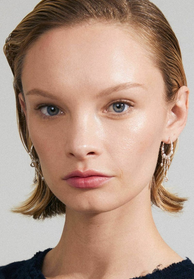 Silver hoop earrings with dangling accents, paired with smooth skin, soft pink lips, and straight, wet-looking hair.