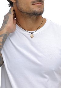 Zwarte koordketting met een kleine beige schildpaddenhanger. De hanger heeft een gestructureerd patroon en hangt om de nek van een persoon die een wit T-shirt draagt.