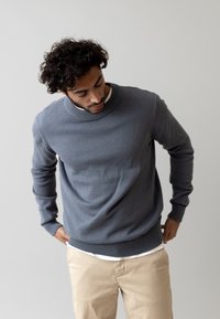 Suéter gris de punto con escote redondo y pu�ños acanalados, llevado sobre una camisa blanca, combinado con pantalones beiges. Textura suave, corte relajado.