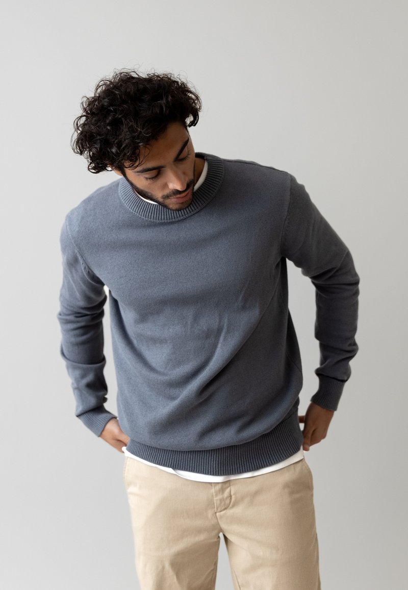 Maglione grigio lavorato a maglia con colletto a girocollo e polsini a costine, indossato sopra una camicia bianca, abbinato a pantaloni beige. Texture morbida, vestibilità rilassata.