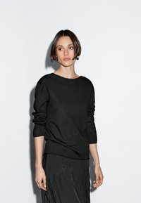 Maglione nero a maniche lunghe con scollo ampio e vestibilità rilassata. Il tessuto sembra morbido con una leggera texture, abbinato a una gonna nera plissettata.