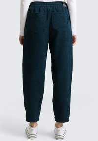 Donkerblauwe corduroybroek met een elastische tailleband, twee achterzakken en een ontspannen pasvorm die naar de enkels taps toeloopt. Witte sneakers zichtbaar.