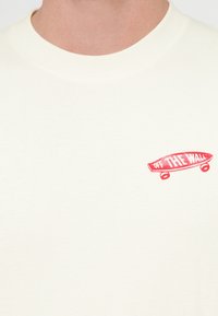 Μπεζ βαμβακερό t-shirt με μικρό κόκκινο γραφικό skateboard και το κείμενο «OFF THE WALL» κοντά στη λαιμόκοψη, στρογγυλή λαιμόκοψη και κοντά μανίκια.