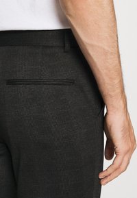 Pantalon à carreaux noirs avec tissu texturé. Comprend une poche arrière et une coupe ajustée. Main posée près de la ceinture.