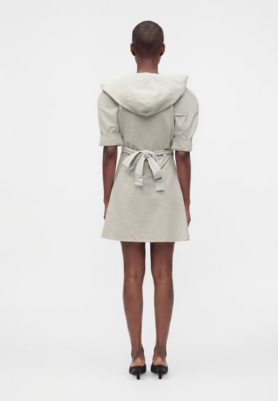 Femme debout de dos, portant une robe courte gris clair avec des manches bouffantes, une ceinture nouée à la taille et une grande capuche, associée à des talons noirs.