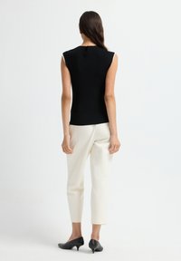 Donna in piedi di spalle, che indossa un top senza maniche nero, pantaloni bianchi alla caviglia e scarpe nere con tacco basso, su uno sfondo bianco.