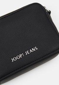 Juodas tekstūruotas odinis piniginė su stačiakampiu pavidalu ir sidabriniu "JOOP! JEANS" logotipu įspaustu priekyje. Užtrauktuko uždarymas viršuje.