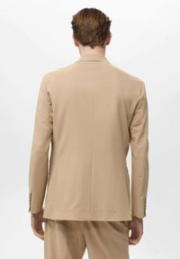 Giacca su misura beige con una texture liscia, spacchetto singolo sul retro e revers standard. Maniche lunghe con dettagli a bottoni.