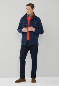 Marinblå vattentät jacka med två framfickor, röd isolerad innerfoder och blå skjorta med knappar, i kombination med mörka jeans och bruna skor.