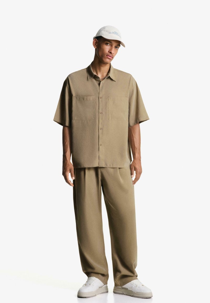 Beige shirt met korte mouwen en twee borstzakken, gecombineerd met bijpassende brede broek. Witte sneakers en een lichte pet maken de outfit compleet.