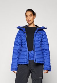 Blå isolert jakke med en puffy design, glidelåslommer og hette. Har en svart innvendig foring og logo på brystet.