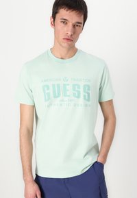 Mint green cotton t-shirt featuring the text "GUESS" in bold, light blue, with a standard crew neck and short sleeves.