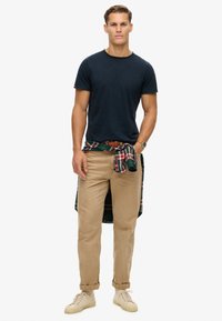 Camiseta de algodón azul marino, chinos caqui y zapatillas beige. Camisa de cuadros verde y roja atada a la cintura. Atuendo sencillo y casual.
