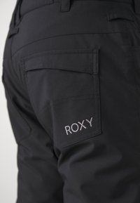 Svart byxor i slitstark tyg har en bakficka och "ROXY"-logotypen broderad i ljusgrått. Närbild av design detaljer.