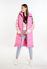 Rosa Steppweste mit Kapuze, ausgestattet mit einem Reißverschluss vorne, gefütterten Taschen und einem gesteppten Design. Kombiniert mit einem gemusterten Pullover und hellblauen Jeans.