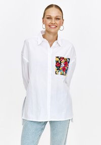 Biała koszula z guzikami, z kolorową kwiatową kieszeni�ą, o luźnym kroju i długich rękawach, zestawiona z jasnoniebieskimi dżinsami.