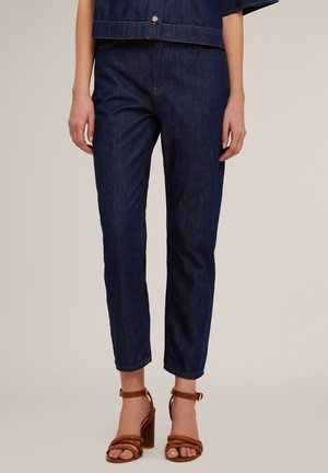 Pantalon en denim bleu foncé avec une coupe slim et des jambes droites, présentant des coutures discrètes et une taille mi-haute. Porté avec des talons bruns à lanières.