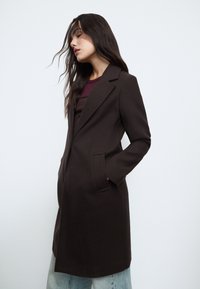 Tmavě hnědý vlněný kabát s jednoručním zapínáním na knoflíky, klopami s výstřihem a bočními kapsami, nošený přes vínový top a světle modré džíny.