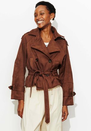 Femme souriante portant une veste en daim marron ceinturée, un pantalon crème et des boucles d'oreilles en forme de cœur, sur fond blanc.