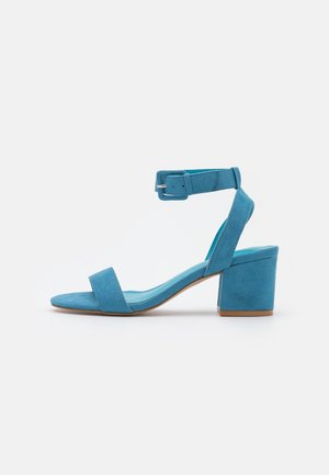 Sandalia de tacón azul de ante con una amplia correa en el tobillo, diseño de punta abierta y tacón cuadrado. Presenta una textura suave y un diseño minimalista.