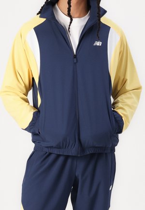 Persoon die een marineblauw en geel New Balance trainingspak draagt, met de handen in de jaszakken, een wit T-shirt eronder en zichtbare gevlochten haarlokken.