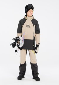 Giacca da neve bicolore in beige e nero con logo, abbinata a pantaloni da neve coordinati. Gli accessori includono guanti, un berretto e occhiali da sci.