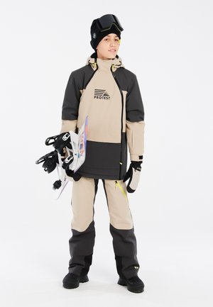Zweifarbige Schneejacke in Beige und Schwarz mit Logo, kombiniert mit passenden Schneehosen. Zubehör umfasst Handschuhe, eine Mütze und eine Skibrille.