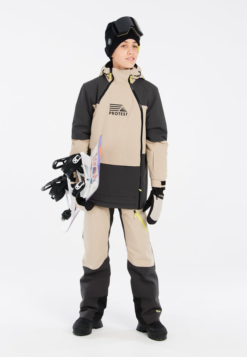 Giacca da neve bicolore in beige e nero con logo, abbinata a pantaloni da neve coordinati. Gli accessori includono guanti, un berretto e occhiali da sci.
