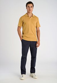 Kortærmet, struktureret polo-shirt i sennepsgul, parret med marineblå bukser og hvide sneakers med farverige detaljer.