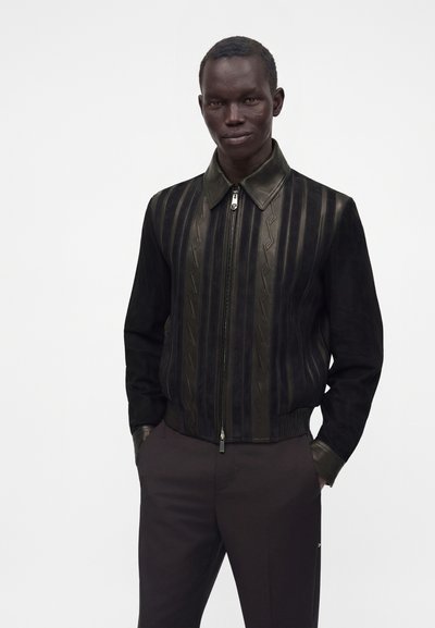 Homme portant une veste noire zippée avec col en cuir et motif rayé, debout les mains dans les poches d'un pantalon sombre sur un fond uni.