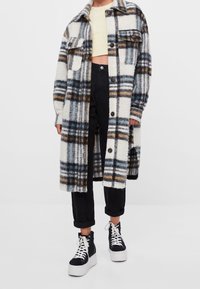 Flauschiger karierter Mantel in Creme mit blauen, schwarzen und braunen Mustern, ausgestattet mit zwei Brusttaschen und schwarzen Knopfverschluss, über dunklen Jeans getragen.