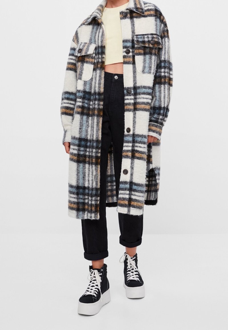 Flauschiger karierter Mantel in Creme mit blauen, schwarzen und braunen Mustern, ausgestattet mit zwei Brusttaschen und schwarzen Knopfverschluss, über dunklen Jeans getragen.