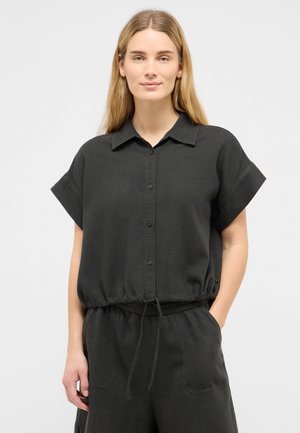 Woman wearing a loose black short-sleeve button-up top with drawstring waist and matching black pants against a plain light background.