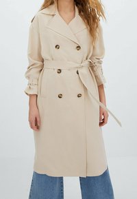 Trench-coat beige à double boutonnage avec de grands boutons, taille ceinturée et manches retroussées, associé à un pantalon en denim à jambes larges.