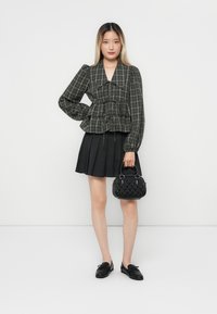 Black and gray plaid peplum top with long sleeves, paired with a pleated black skirt and a black textured handbag. Black shoes complete the look.